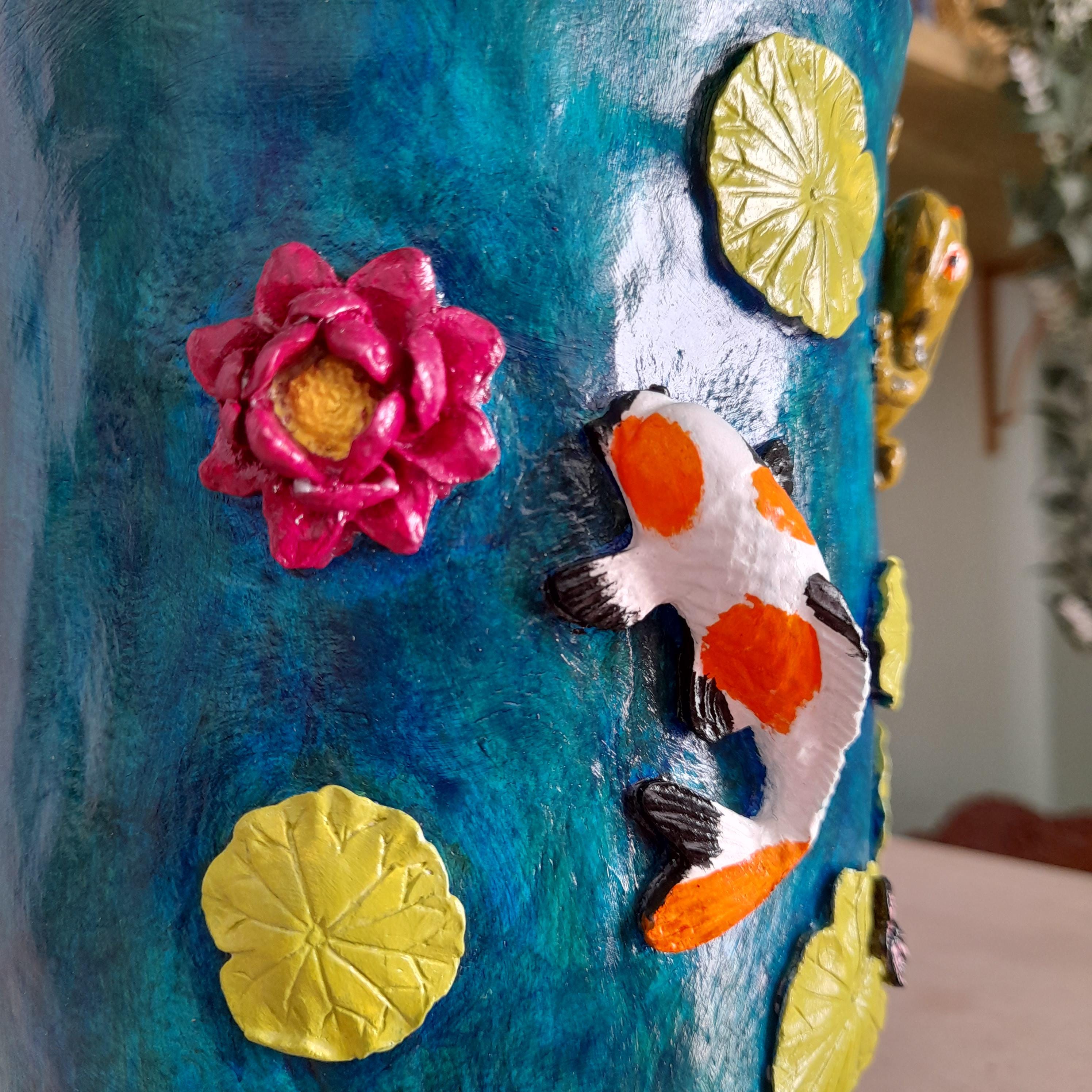 Detalhe de uma superfície cilíndrica decorativa com fundo texturizado azul e elementos 3D em relevo, incluindo uma flor de lótus rosa com centro amarelo, nenúfares amarelos e um peixe koi branco com marcas laranja e pretas.