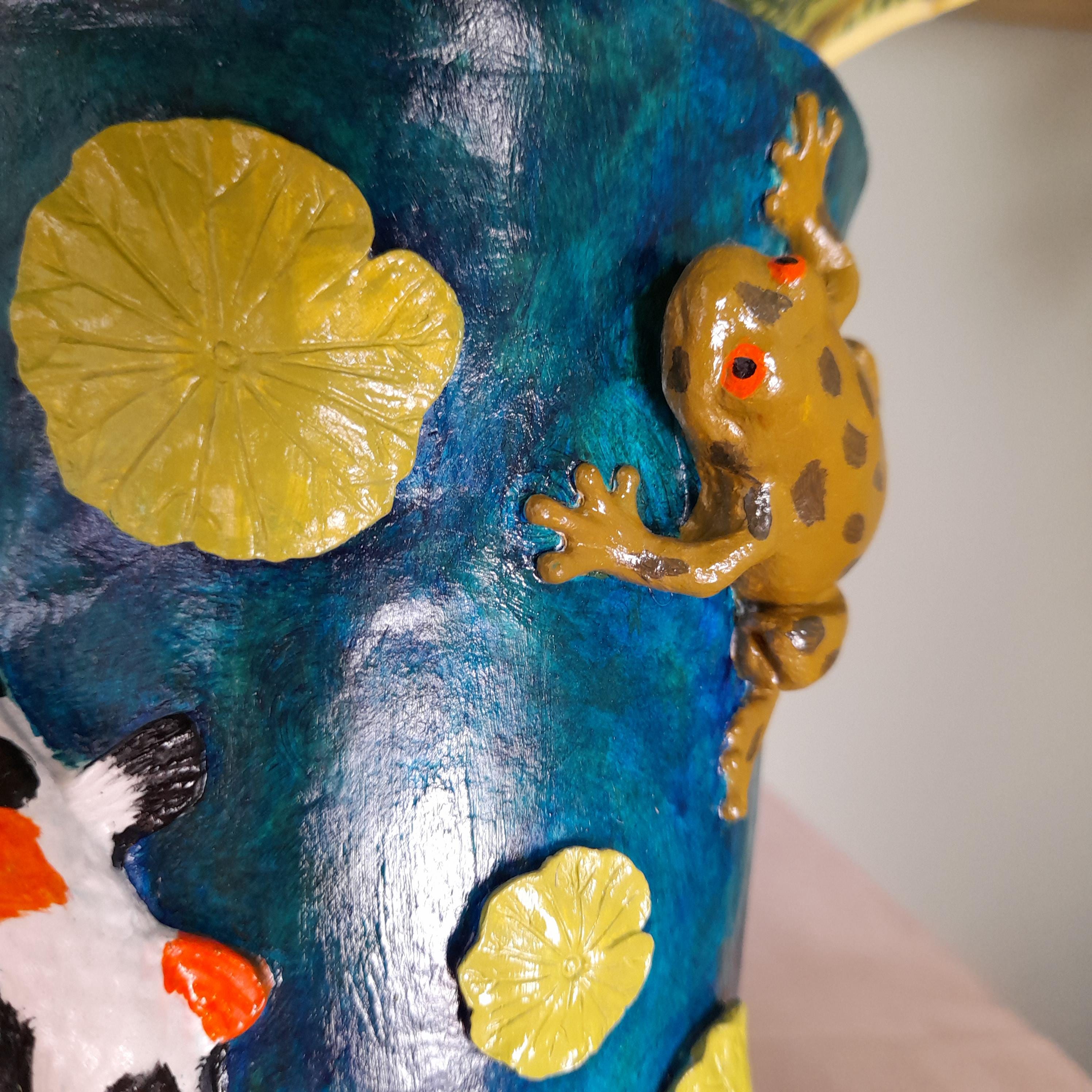 Detalhe de um vaso decorativo pintado à mão, com fundo estilo aguarela em azul, nenúfares amarelos, uma rã castanha a subir pela lateral, e parte de uma ilustração de peixe koi.