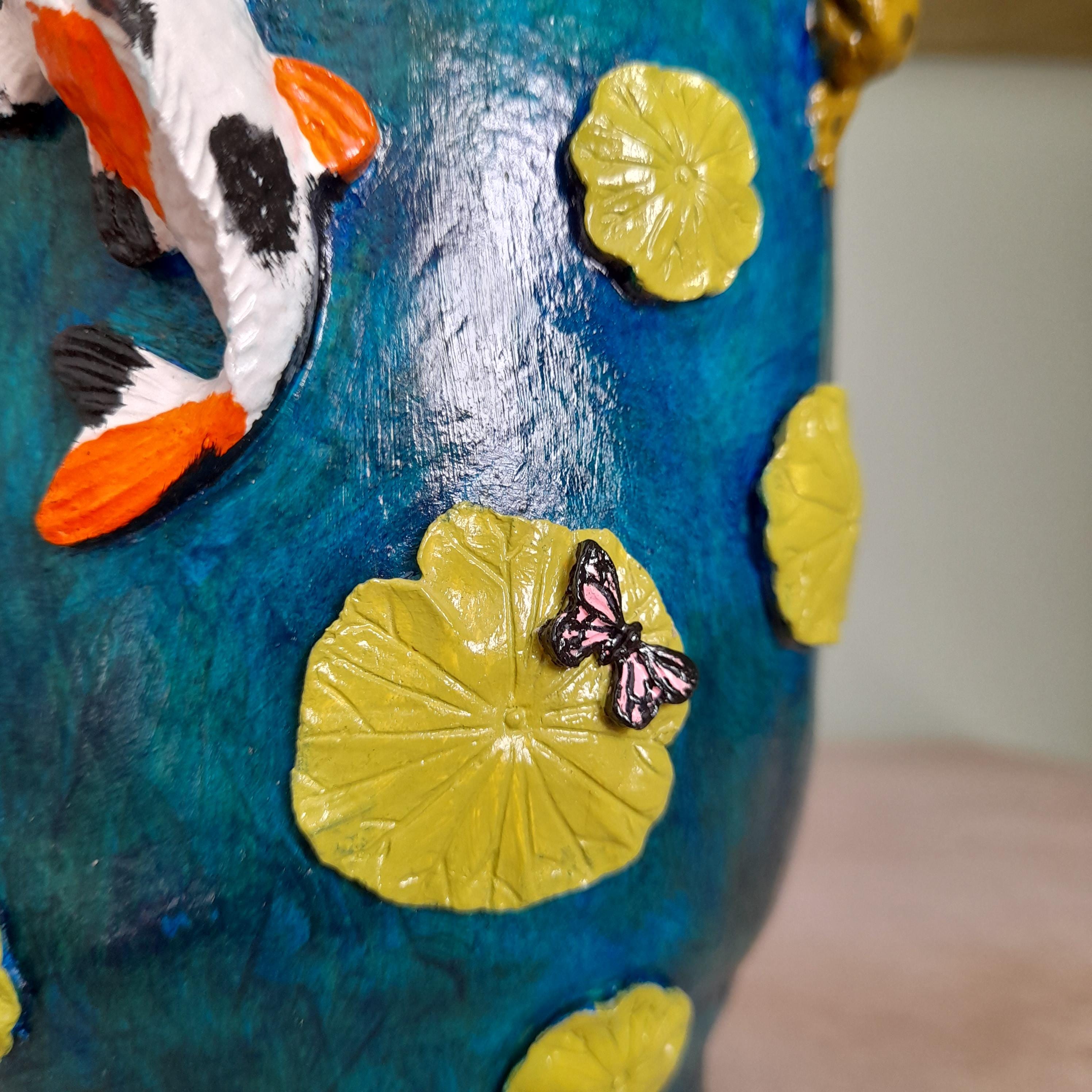 Detalhe de um vaso decorativo de cerâmica azul com nenúfares amarelos, um peixe koi e um enfeite de borboleta.