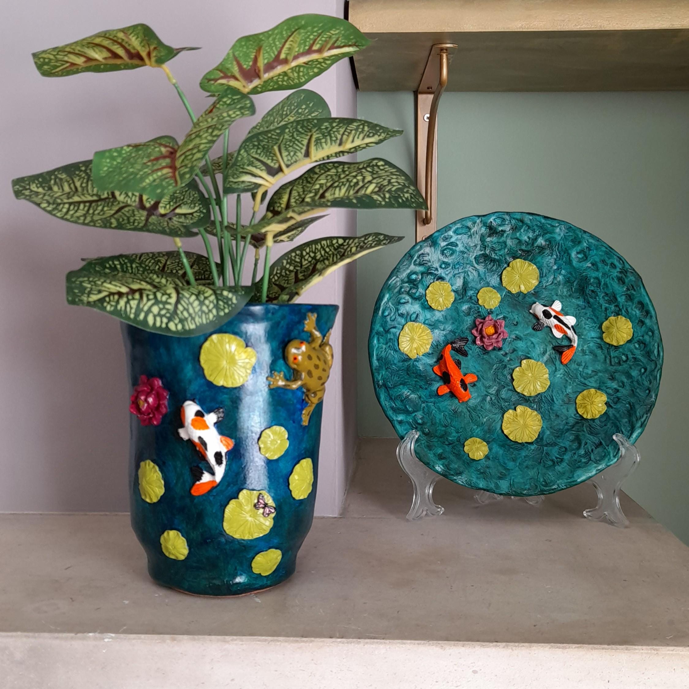 Um vaso decorativo de cerâmica verde-azulado com nenúfares amarelos e peixes koi, com uma planta de folhas verdes, exposto junto a um prato decorativo a condizer sobre suportes transparentes.