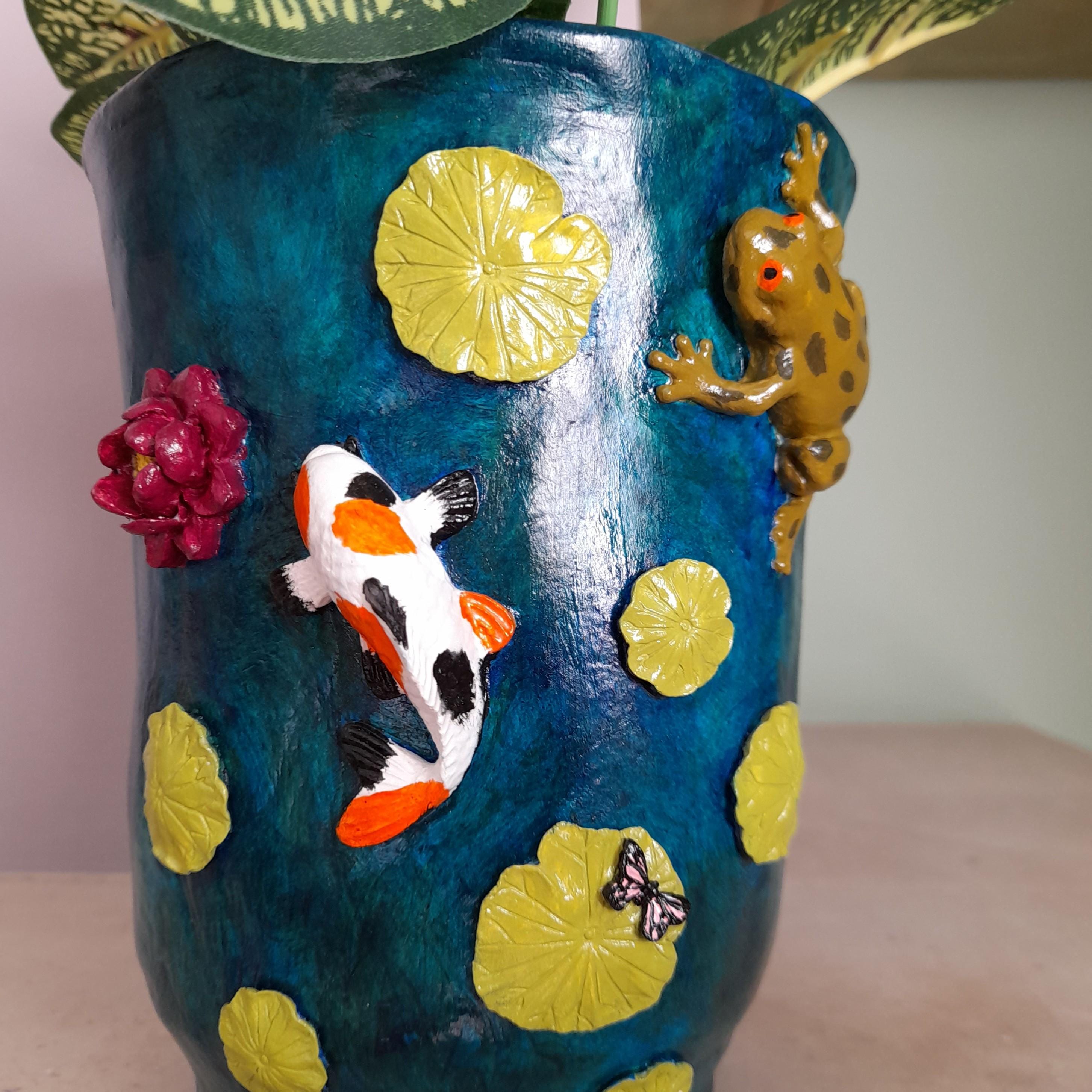 Um vaso decorativo de cerâmica com fundo texturizado azul-escuro, apresentando elementos 3D em relevo de um peixe koi, uma rã, folhas de nenúfar, uma flor e uma borboleta, com folhas verdes na parte superior.